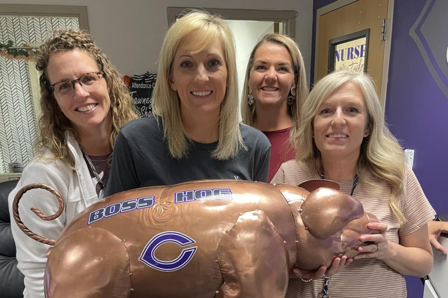 The four front office staff ladies are seen holding the large, copper pig with the Canyon High C logo facing towards the camera. On the right is Linda Thompson wearing a pinkish shirt with white wavey hair, next to her on the left is Tawny Shawltry wearing a red shirt with brown hair and blonde high lights, next to her on her left is Magan Doak with blond straight hair wearing a gray shirt. On the far left is Nurse, Paige Tucker with curly light brown hair wearing a white jacket.